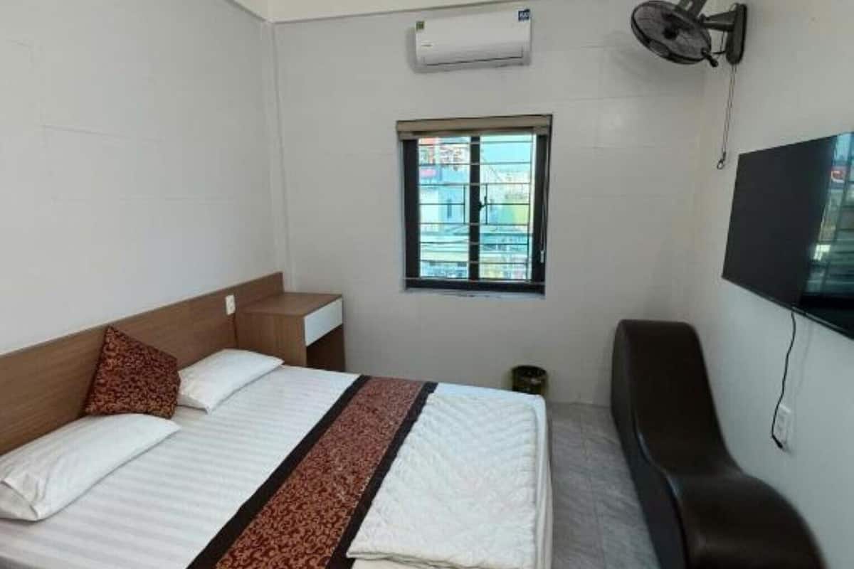Quarto de hotel com cama, televisão, janela e ventoinha fixada na parede.