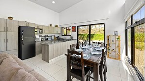A modern kitchen with a central island, dining area with a glass table, and large windows offering a view of the outdoors. - Family Getaway with Private Pool | 15 Mins to Beach & Mount (Veintisiete de Abril, Guanacaste)