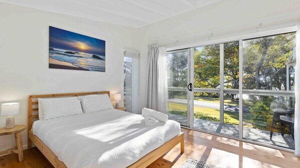 A bedroom with a wooden bed, a nightstand, a painting of a sunset, and a sliding glass door leading to an outdoor area with a table and chairs. - Bendalong Beachside near Washerwomans Beach (Bendalong)