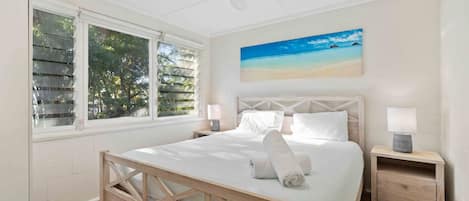 A bedroom with a bed, two bedside tables, a ceiling fan, a window with blinds, and a beach painting on the wall.