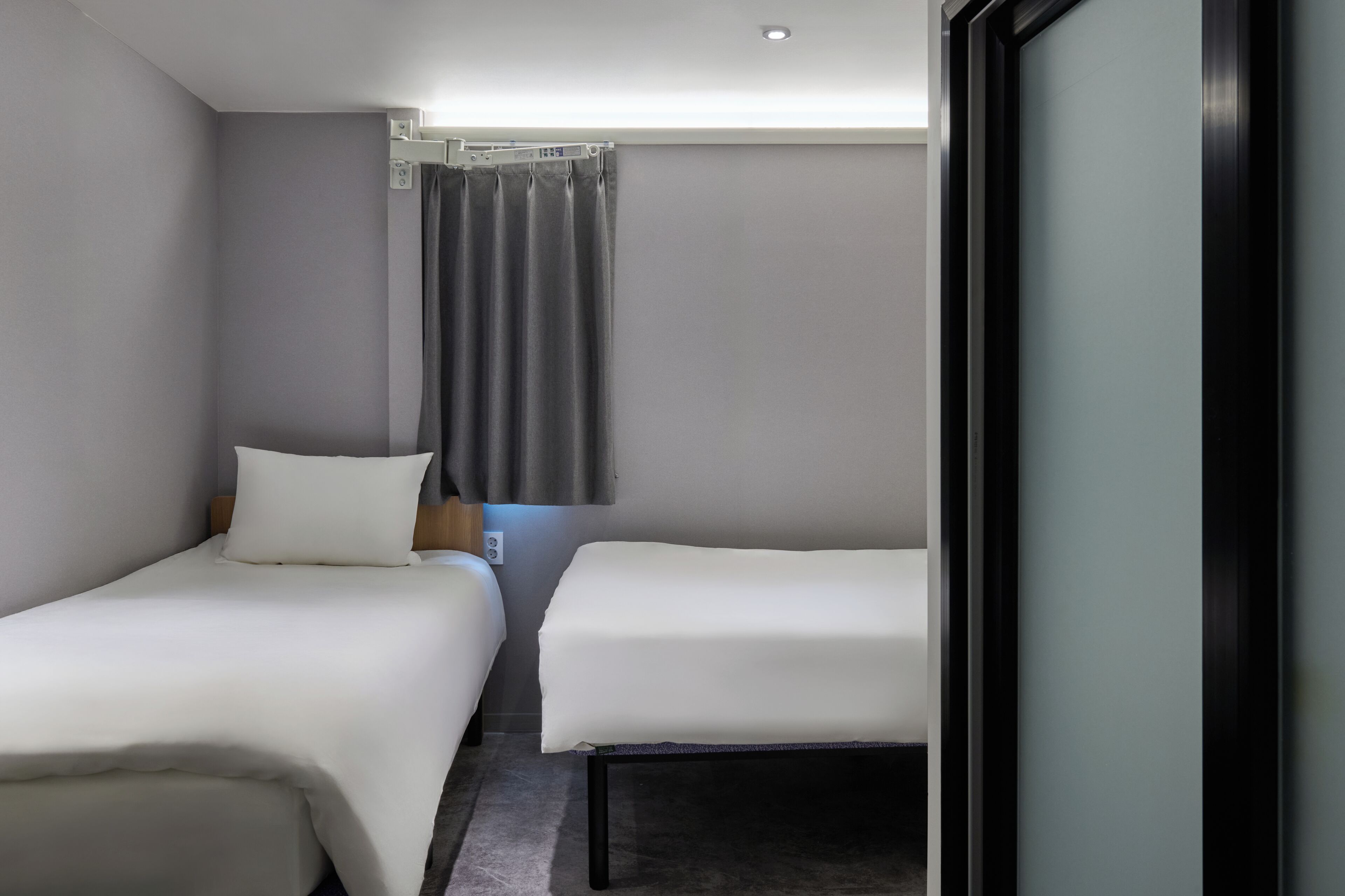A hotel room with two single beds, a small table, a chair, a window with curtains, and a door.