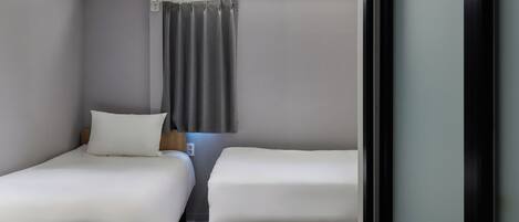 A hotel room with two single beds, a small table, a chair, a window with curtains, and a door.