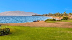 A coastal view with a beach, a mountain in the background, and a grassy area with a bench. - Stunning 2BR condo in South Kihei with ocean views! (Kihei)