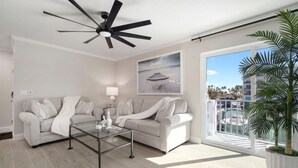 Un salon moderne doté d’un ventilateur de plafond, d’une table basse en verre, d’un canapé et d’une grande fenêtre offrant une vue sur des palmiers et une plage.