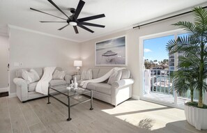 Ein modernes Wohnzimmer mit Deckenventilator, Glastisch, Sofa und großem Fenster mit Blick auf Palmen und einen Strand.