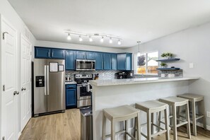 A modern kitchen with blue cabinets, stainless steel appliances, and a bar area with stools. - Modern Home | Game Room, Putt Putt & Pets Welcome! (Colorado Springs)