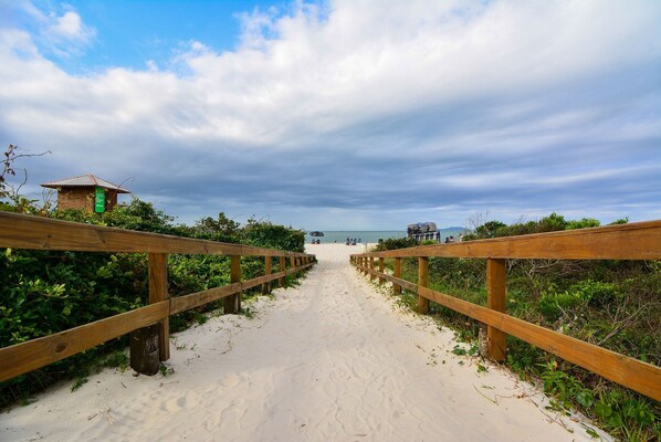 A wooden boardwalk leading to a beach with a small structure on the left. - IL CAMPANÁRIO (Florianópolis)