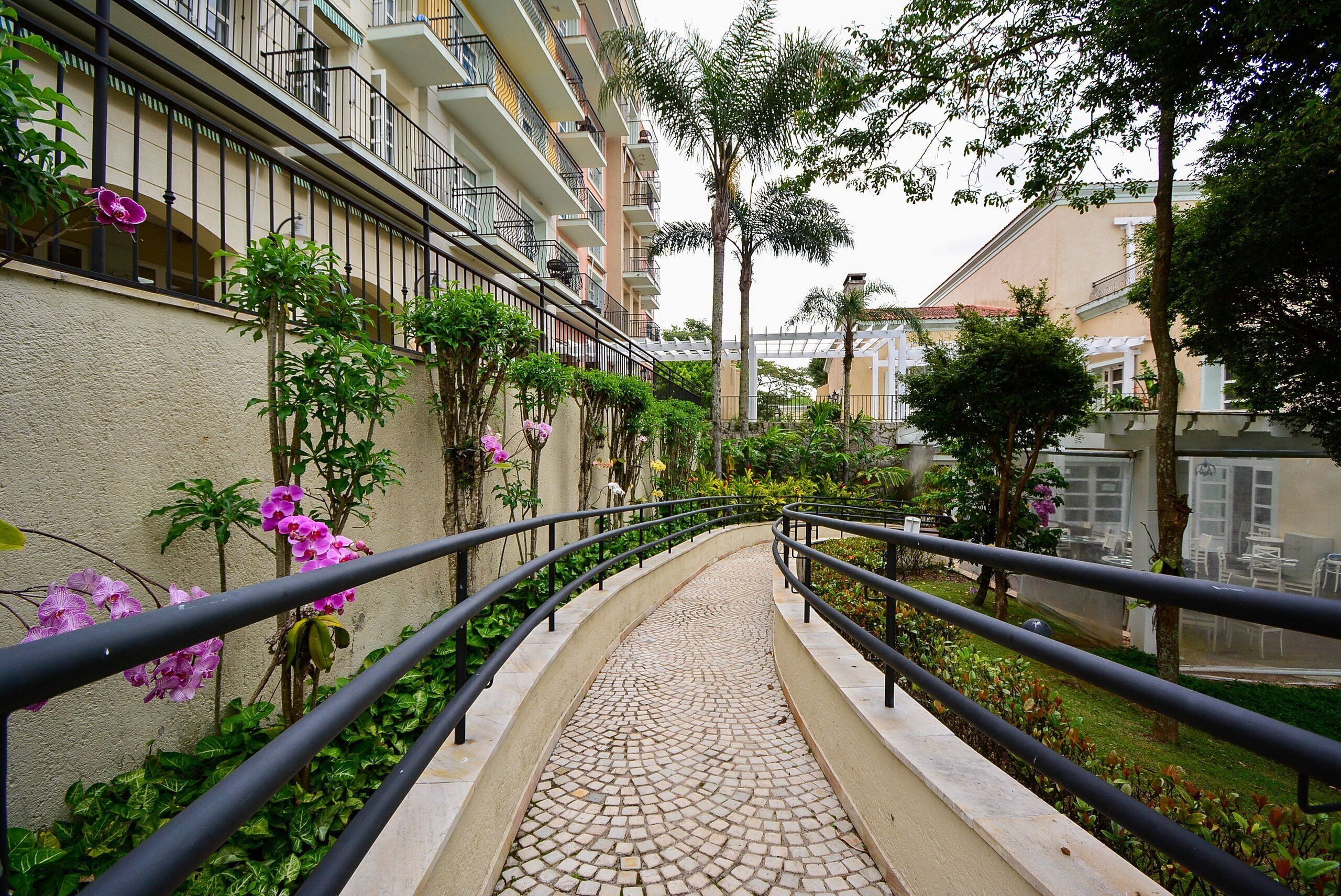 A pathway with a tiled surface leading to a building with balconies and a garden area with various plants and flowers.