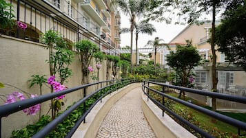 A pathway with a tiled surface leading to a building with balconies and a garden area with various plants and flowers.