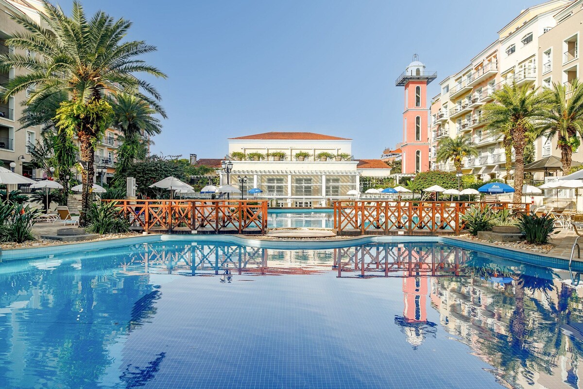 A swimming pool with a clear blue water surface, surrounded by palm trees and a building with a red tower.