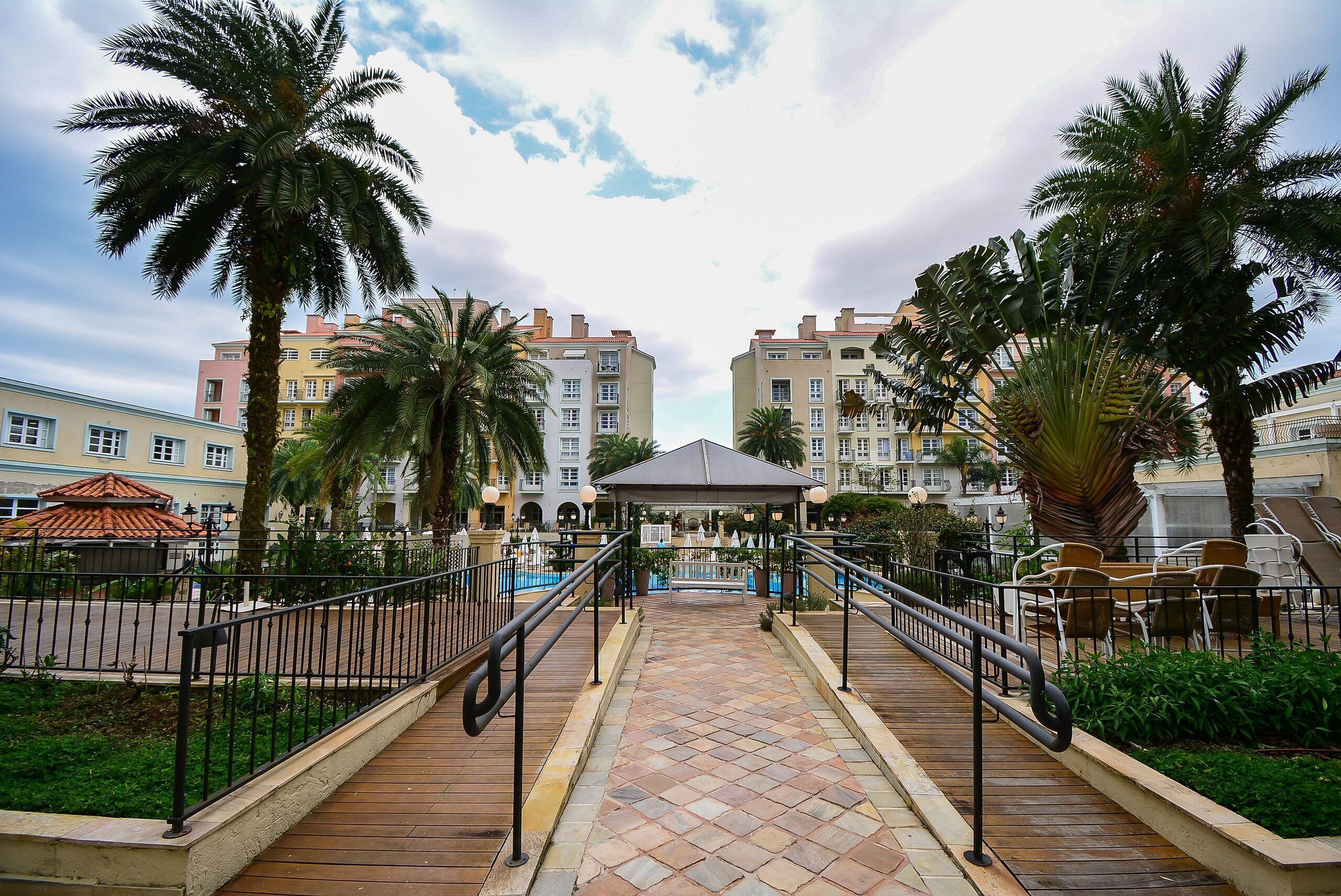 Un sendero conduce a un pabellón rodeado de palmeras y una zona de piscina, con edificios de varios pisos al fondo.
