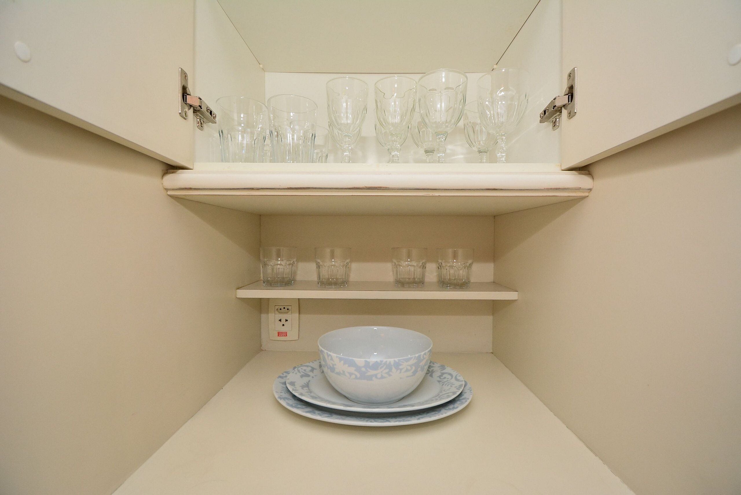A kitchen with a white countertop, a white shelf above holding clear glassware, and a white bowl with a blue pattern on a matching plate below.