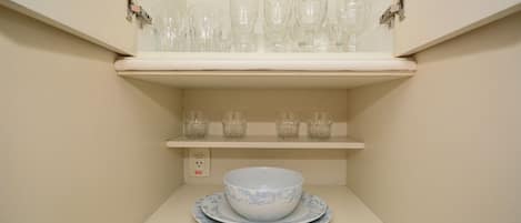 A kitchen with a white countertop, a white shelf above holding clear glassware, and a white bowl with a blue pattern on a matching plate below.