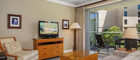 Habitación de hotel con televisión, muebles de madera, sofá, lámpara y balcón con mobiliario exterior.