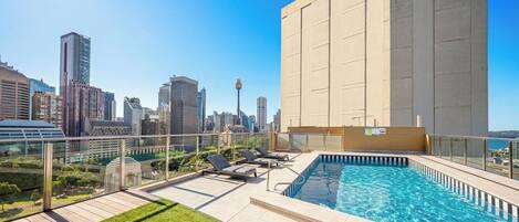 A rooftop pool with lounge chairs, a city skyline view, and a large building in the background.