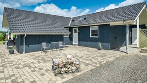 A two-story building with a grey tiled roof, a covered patio area with outdoor furniture, and a pile of firewood. - Cozy home in Ansager with sauna (Ansager)