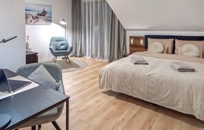 Een moderne hotelkamer met een houten vloer, een bed met een beige deken, een bureau met een laptop, een blauwe fauteuil en een schilderij van een strandgezicht.