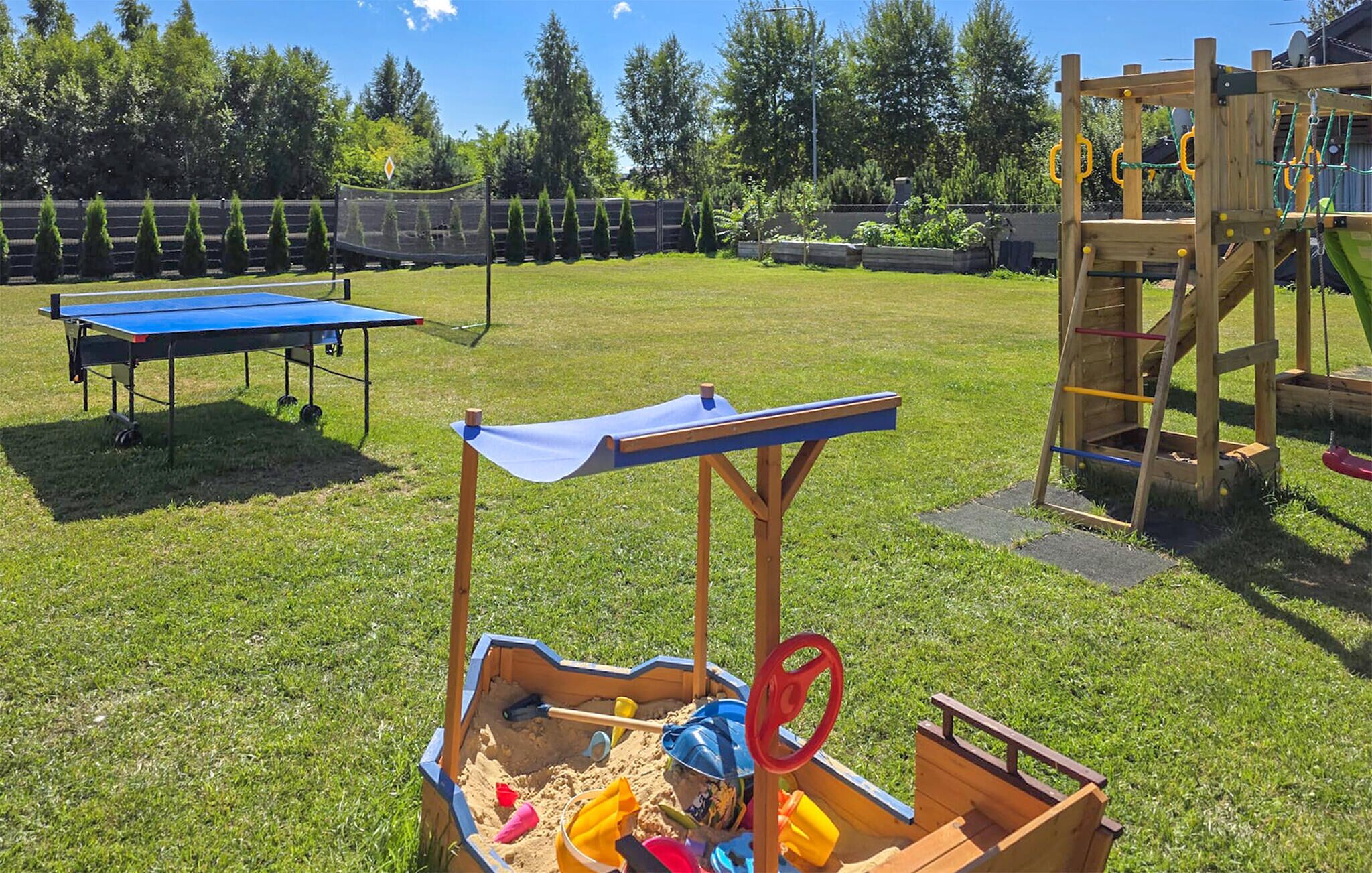 Un jardin avec un bac à sable, une table de ping-pong et une structure de jeu en bois.