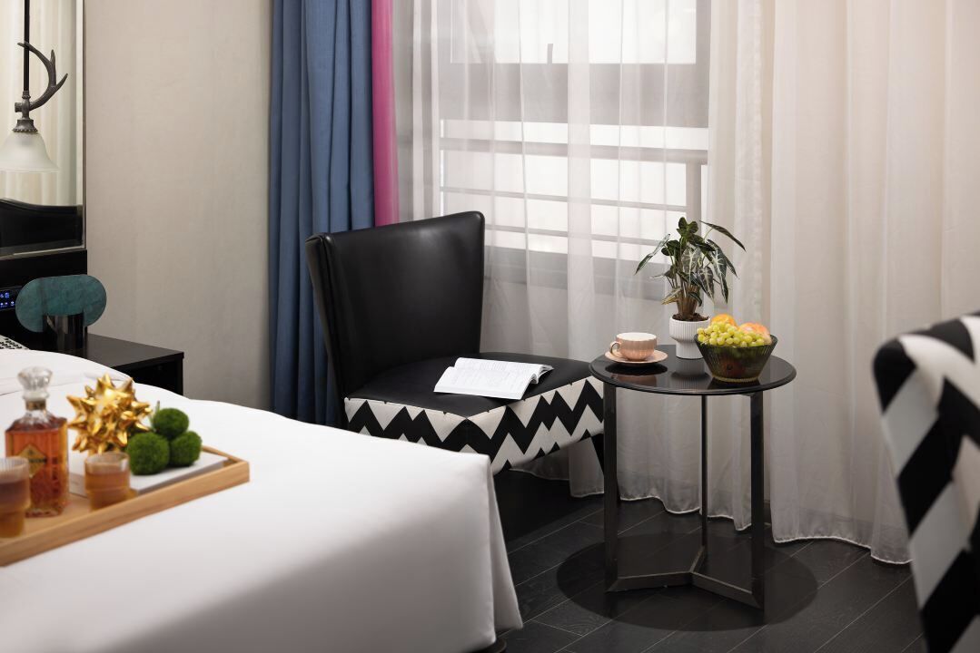 A hotel room with a bed, a black leather chair, a small round table with a plant and fruit bowl, a tray with drinks and a decorative item, and a window with sheer curtains.