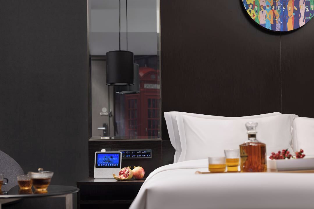A modern hotel room with a bed, bedside table, a digital clock, a pomegranate, a bottle, and a glass of amber liquid.
