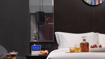 A modern hotel room with a bed, bedside table, a digital clock, a pomegranate, a bottle, and a glass of amber liquid.