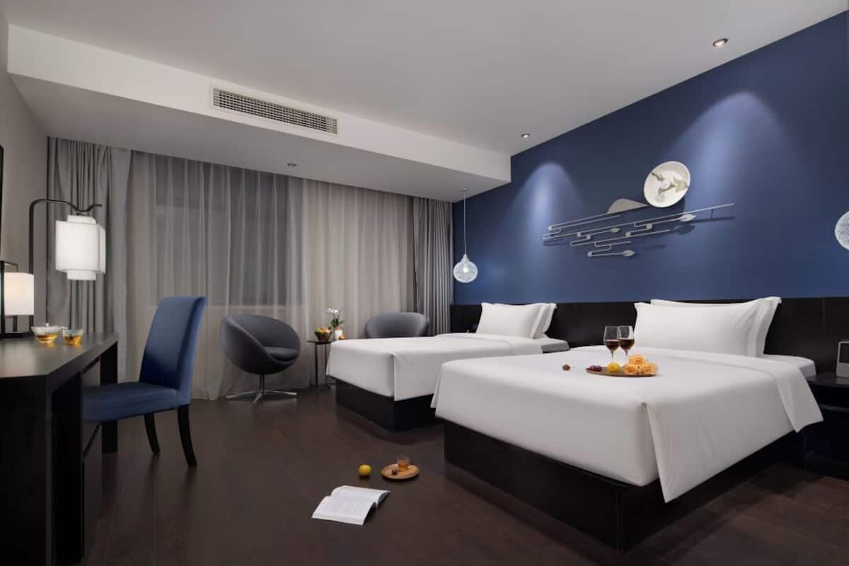 Une chambre d’hôtel avec deux lits, un bureau, une chaise, une table à manger avec un plateau de nourriture et de boissons, et un élément décoratif mural.