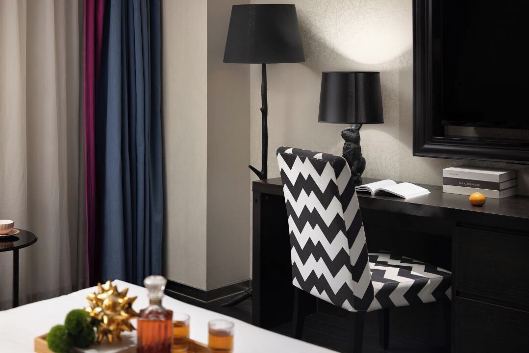 A hotel room with a black and white zigzag patterned chair, a black floor lamp, a flat-screen TV, and a small table with a bottle and glasses.