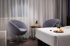 Une chambre d’hôtel avec deux fauteuils gris, une petite table avec des fruits et des boissons, et un lit recouvert d’une couverture blanche.