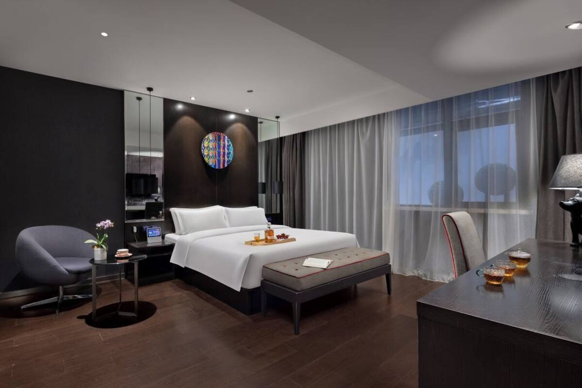 Une chambre d’hôtel moderne avec un grand lit, un bureau, une chaise, une petite table avec une plante et une grande fenêtre avec des rideaux.
