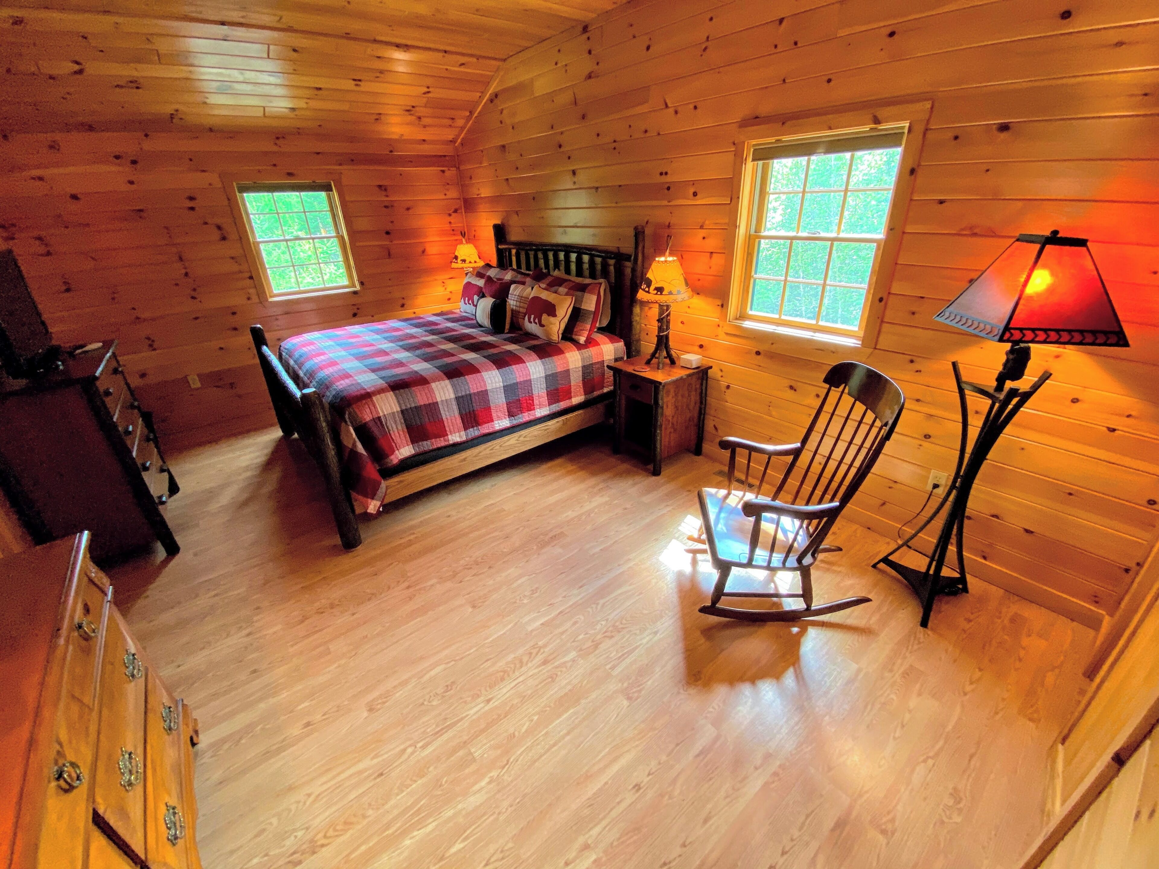 Une pièce chaleureuse dans un chalet, avec un plancher en bois, un lit recouvert d’une couverture à carreaux, un fauteuil à bascule et deux fenêtres.