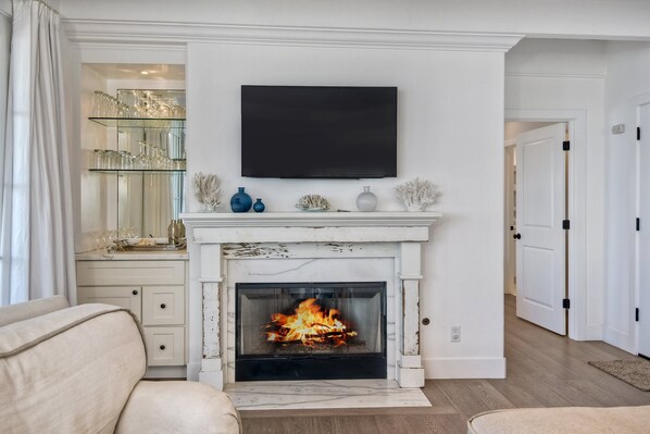 A living room with a fireplace, a television mounted above it, and a glass shelving unit with decorative items.