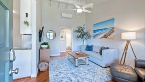 A modern living room with a white ceiling fan, a large painting of a beach, a white sofa, a coffee table, and a leather armchair. - 3080 Lincoln - Unit 12 (Carlsbad)