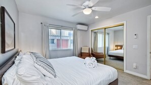 A hotel room with a bed, a ceiling fan, a window with curtains, a chair, and a mirror. - 3080 Lincoln - Unit 12 (Carlsbad)