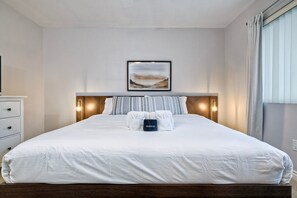 A neatly made bed with white linens and a folded towel with a "Makeup" label, a framed landscape painting above it, and a bedside lamp on the nightstand. - 3080 Lincoln - Unit 12 (Carlsbad)