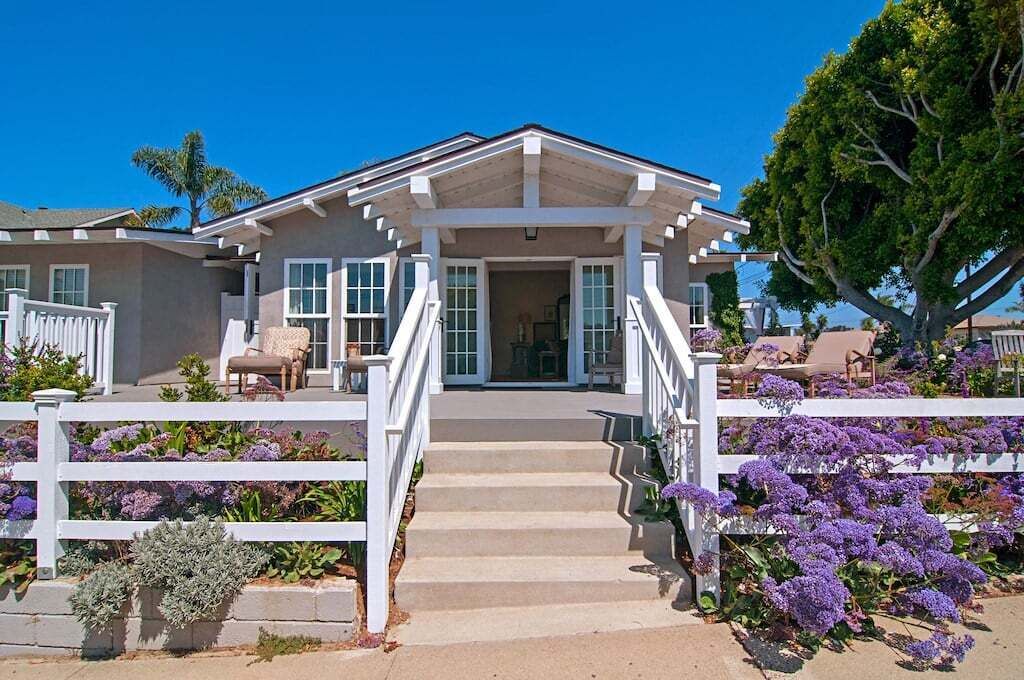 A two-story house with a covered front porch, a staircase leading to the entrance, and a garden with purple flowers.
