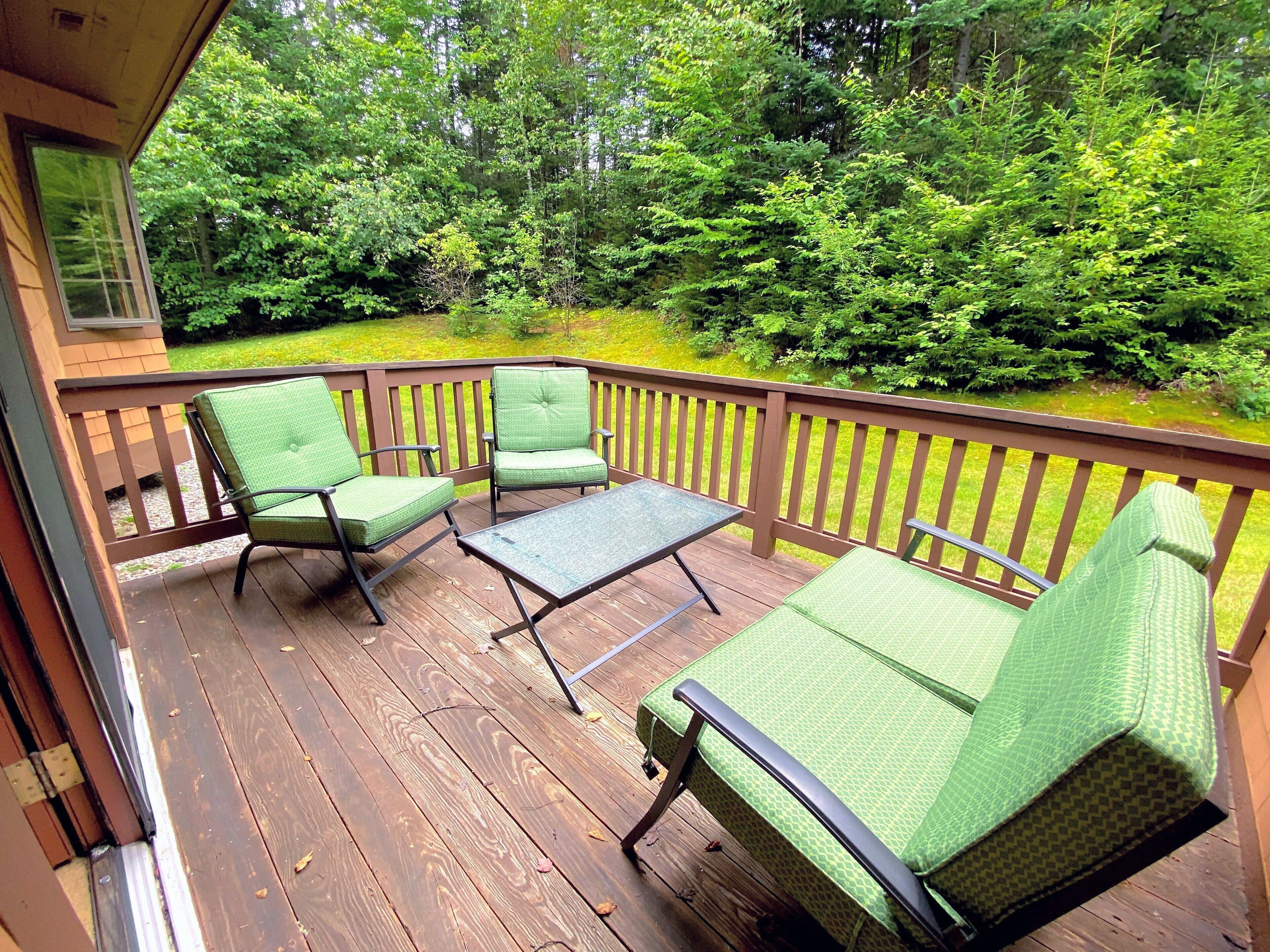 A wooden deck with green cushioned chairs and a small table, surrounded by dense greenery.