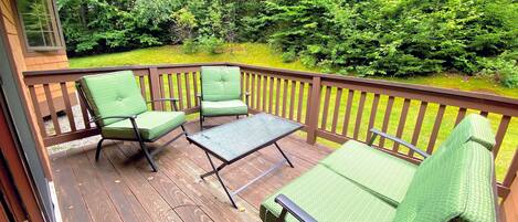 A wooden deck with green cushioned chairs and a small table, surrounded by dense greenery.
