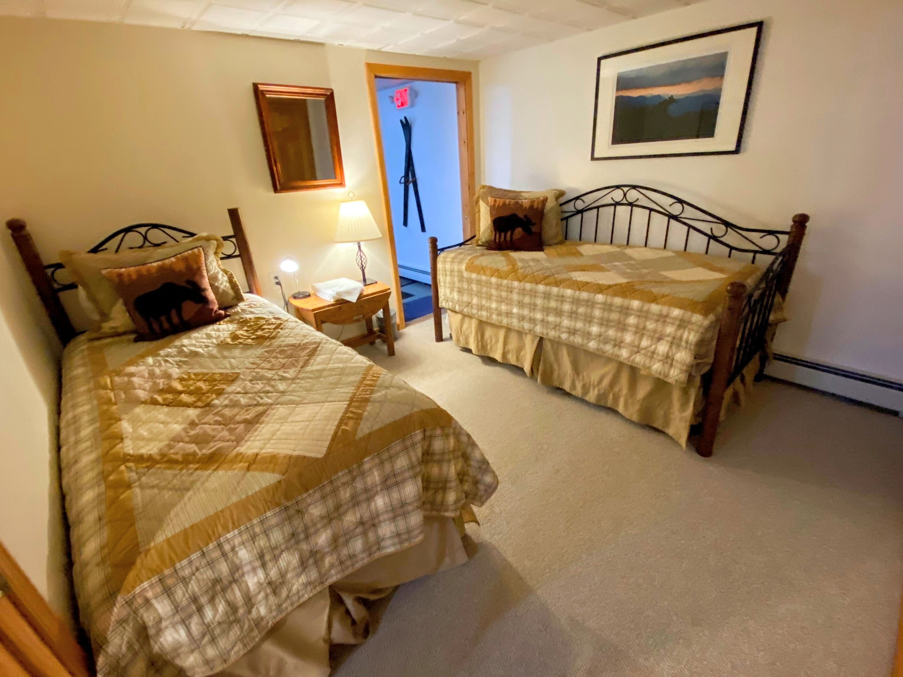 Quarto com duas camas de solteiro, mesa de cabeceira com abajur, porta com câmera de segurança e quadro na parede.