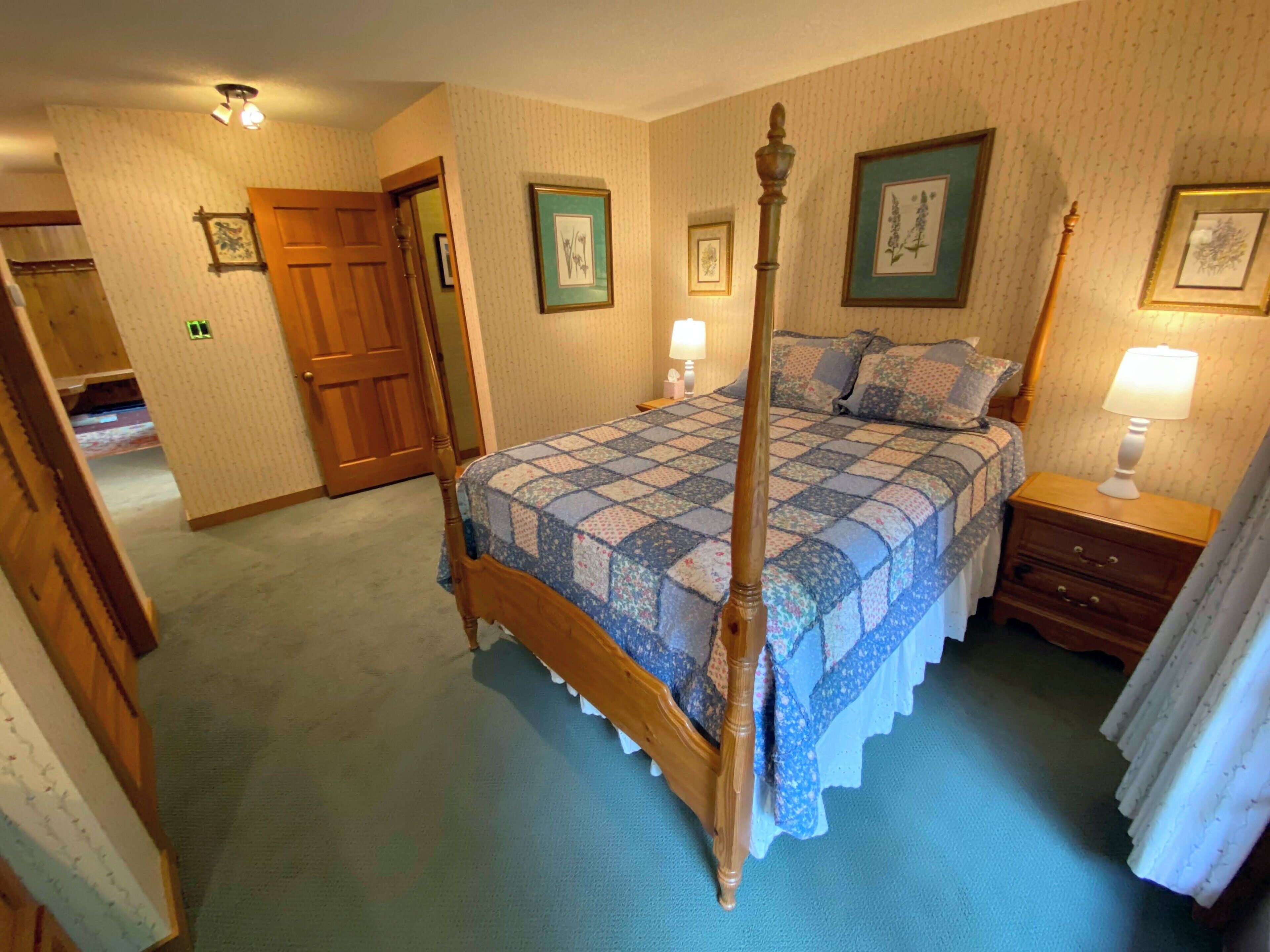 Une chambre à coucher avec un lit à baldaquin, une table de chevet en bois, une lampe et des œuvres d’art encadrées accrochées aux murs.