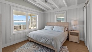 Ein Schlafzimmer mit einem großen Bett, einem Deckenventilator, einem Nachttisch mit Lampe und Blick auf den Strand durch ein Fenster.
