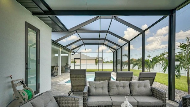 A covered patio with wicker furniture, a glass table, and a view of a swimming pool and greenery. - 290) Perfect for Families! (Cape Coral)