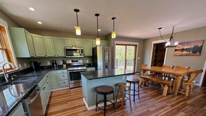 A spacious kitchen with green cabinets, a large island with stools, a wooden dining table, and a view of the outdoors through a sliding door. - 1800ml - As Seen on TV 1 Mile From Skiing, Great Views, hot Tub, Fire Pit, Beach! Pool gym Passes (Bretton Woods)