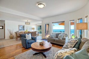 A spacious living room with a blue sofa, a grey sofa, and a round coffee table. There is a dining area with a table and chairs, and a balcony with a view of the ocean. - 2751 Ocean Street (Carlsbad)