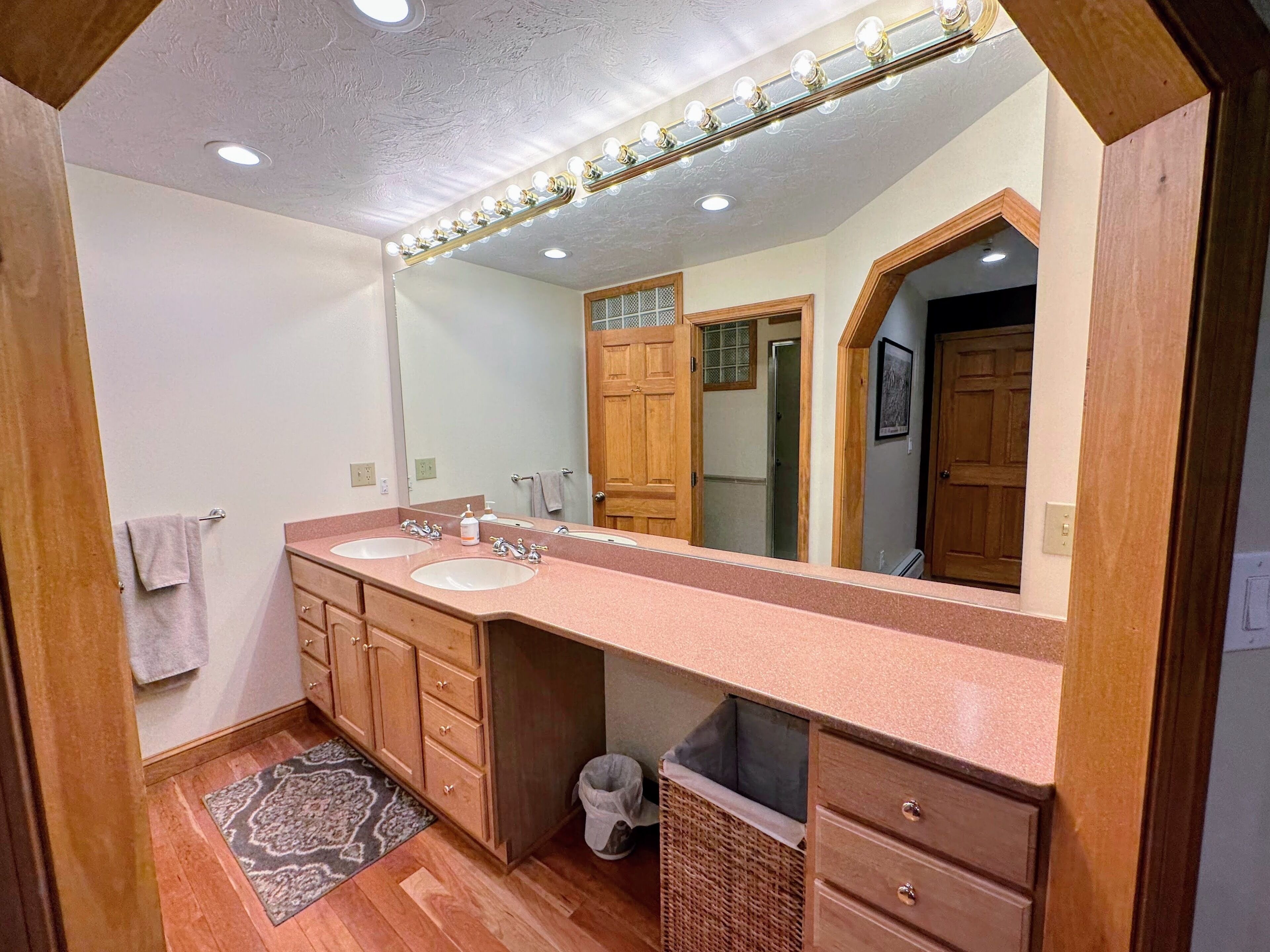 Un bagno con due lavabi, mobili impiallacciati in legno, un grande specchio illuminato, un tappeto e un cestino.