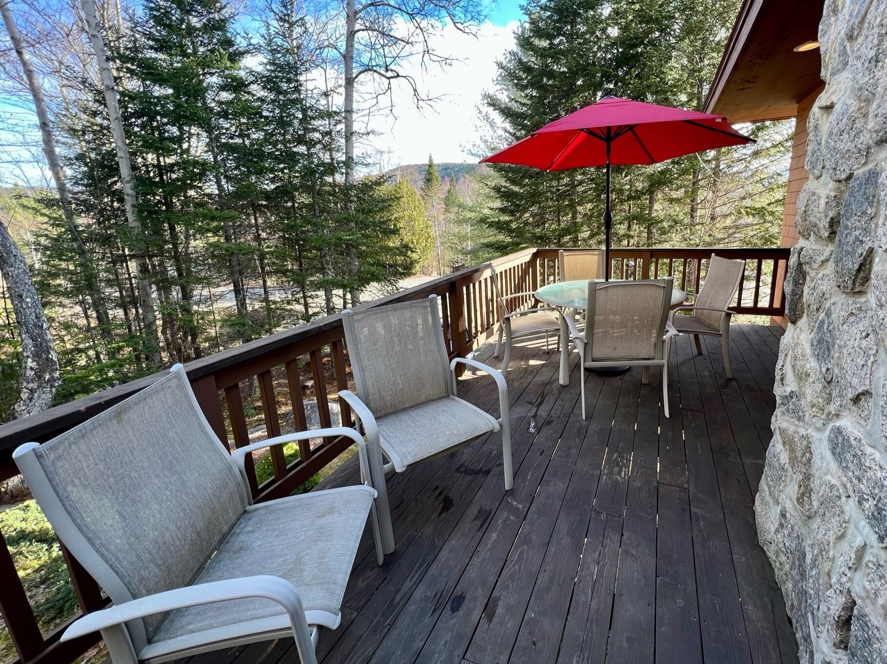 A wooden deck with two white chairs and a table, a red umbrella, and a stone wall.