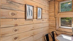 A wooden cabin interior with two framed fern pictures on the wall, a window with a view of greenery, and a table with chairs. - Amazonia (Rogers)