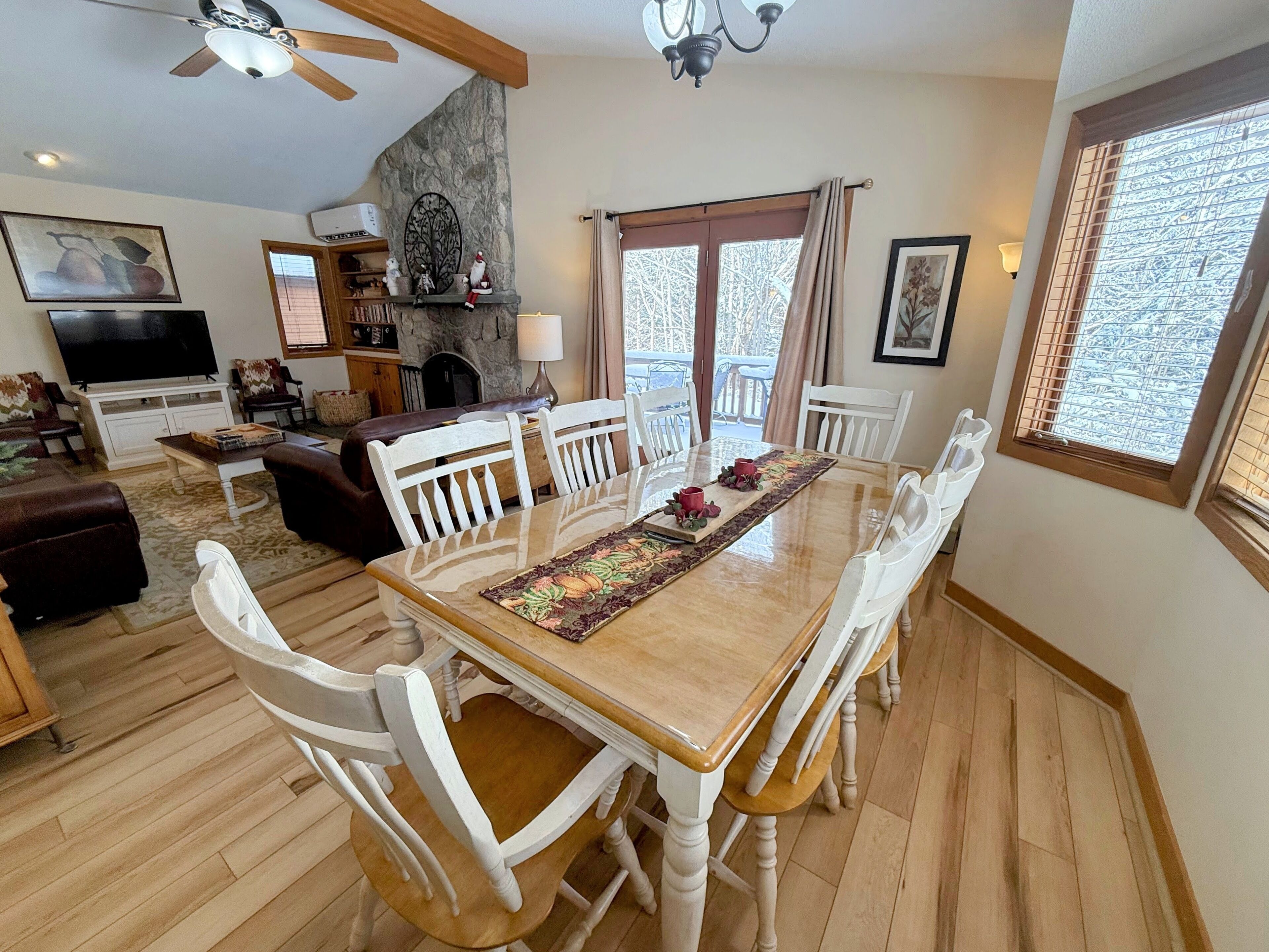 Un comedor con mesa de madera y sillas blancas, chimenea y televisor en la sala.