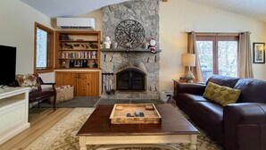 A living room with a stone fireplace, a bookshelf, a television, and a leather sofa. - Fc14 Slopeside Bretton Woods Cottage With AC, Large Patio and Private Yard! Walk to Slopes! (Bretton Woods)