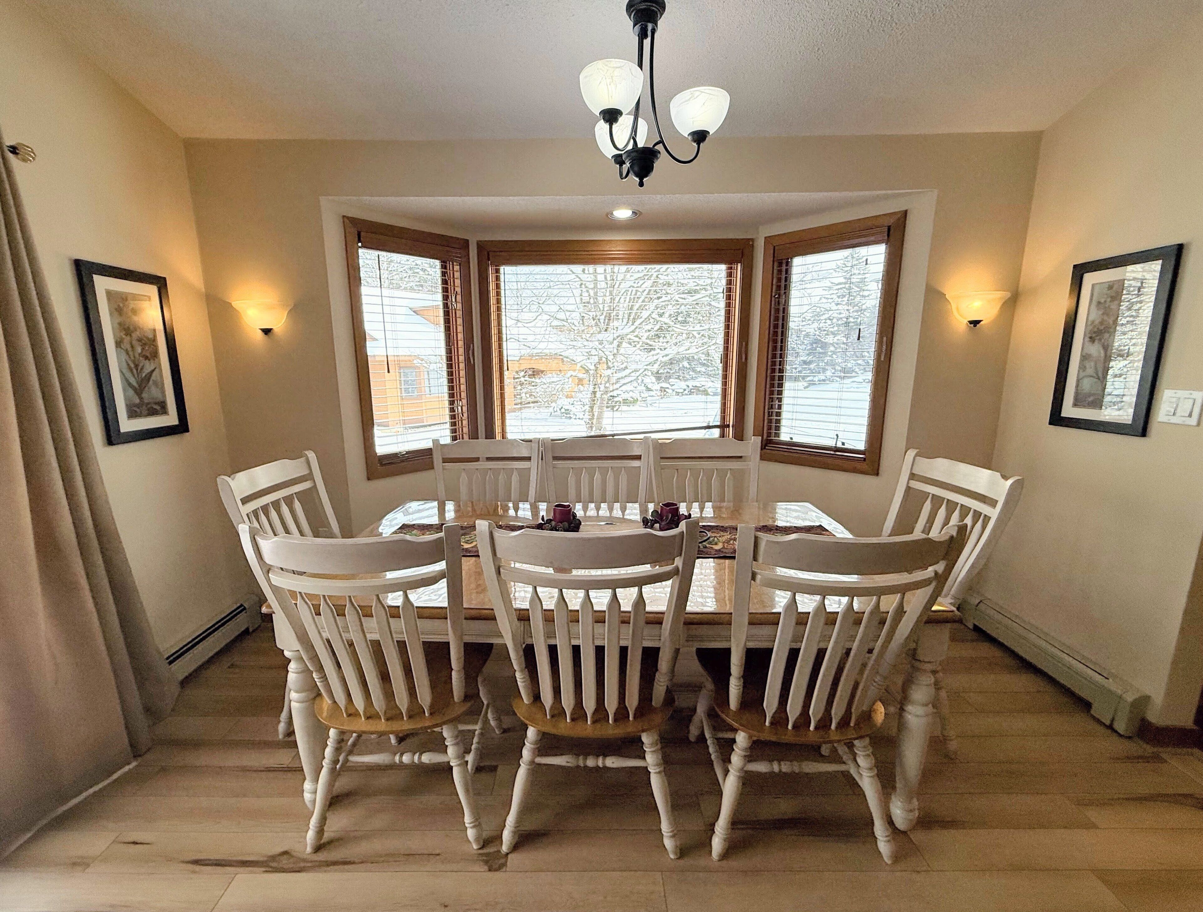 A dining area with a round table and six chairs, large windows with blinds, and wall-mounted light fixtures.