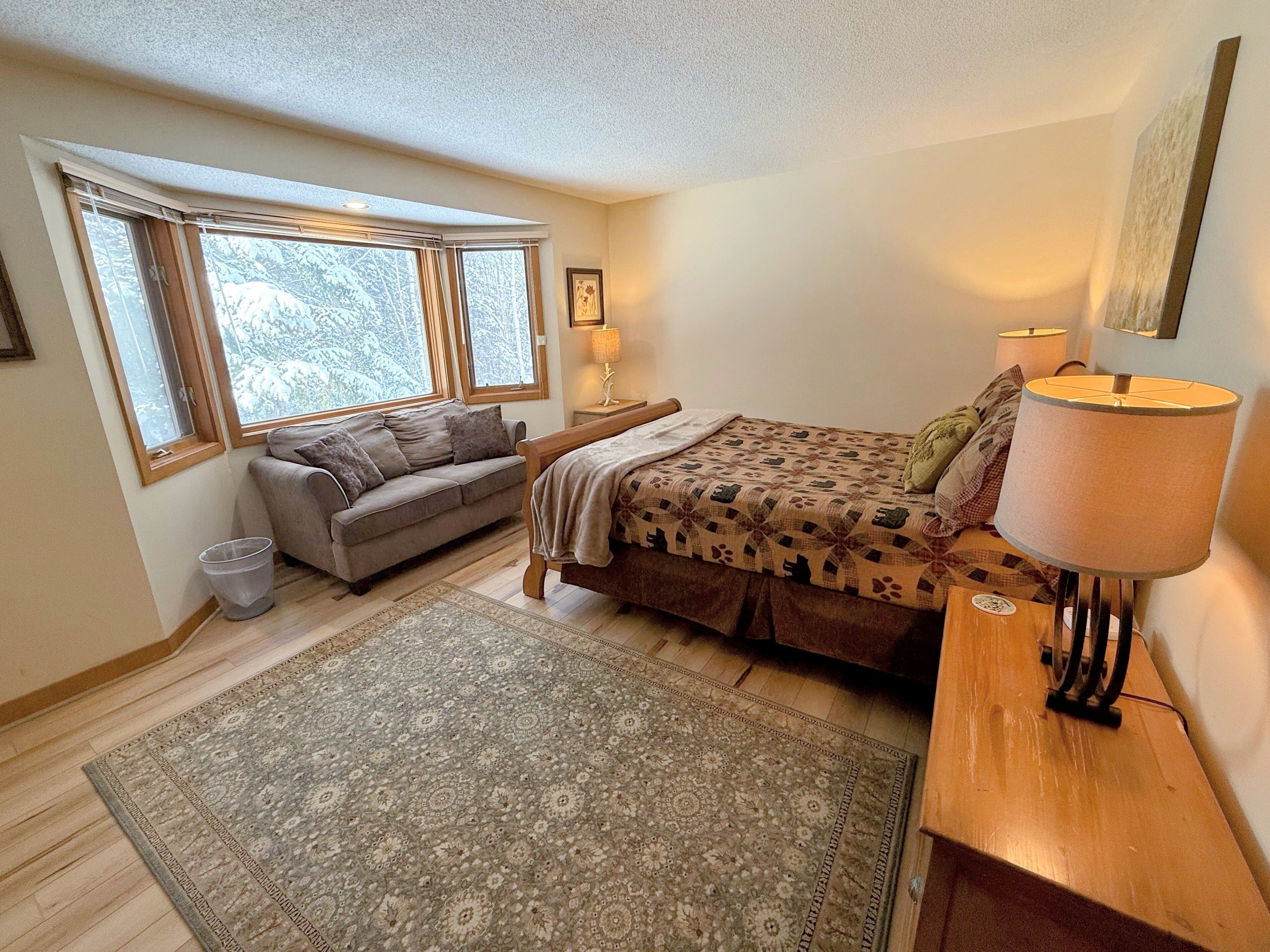 A bedroom with a bed, a sofa, a lamp, a rug, and a window with a view of trees.